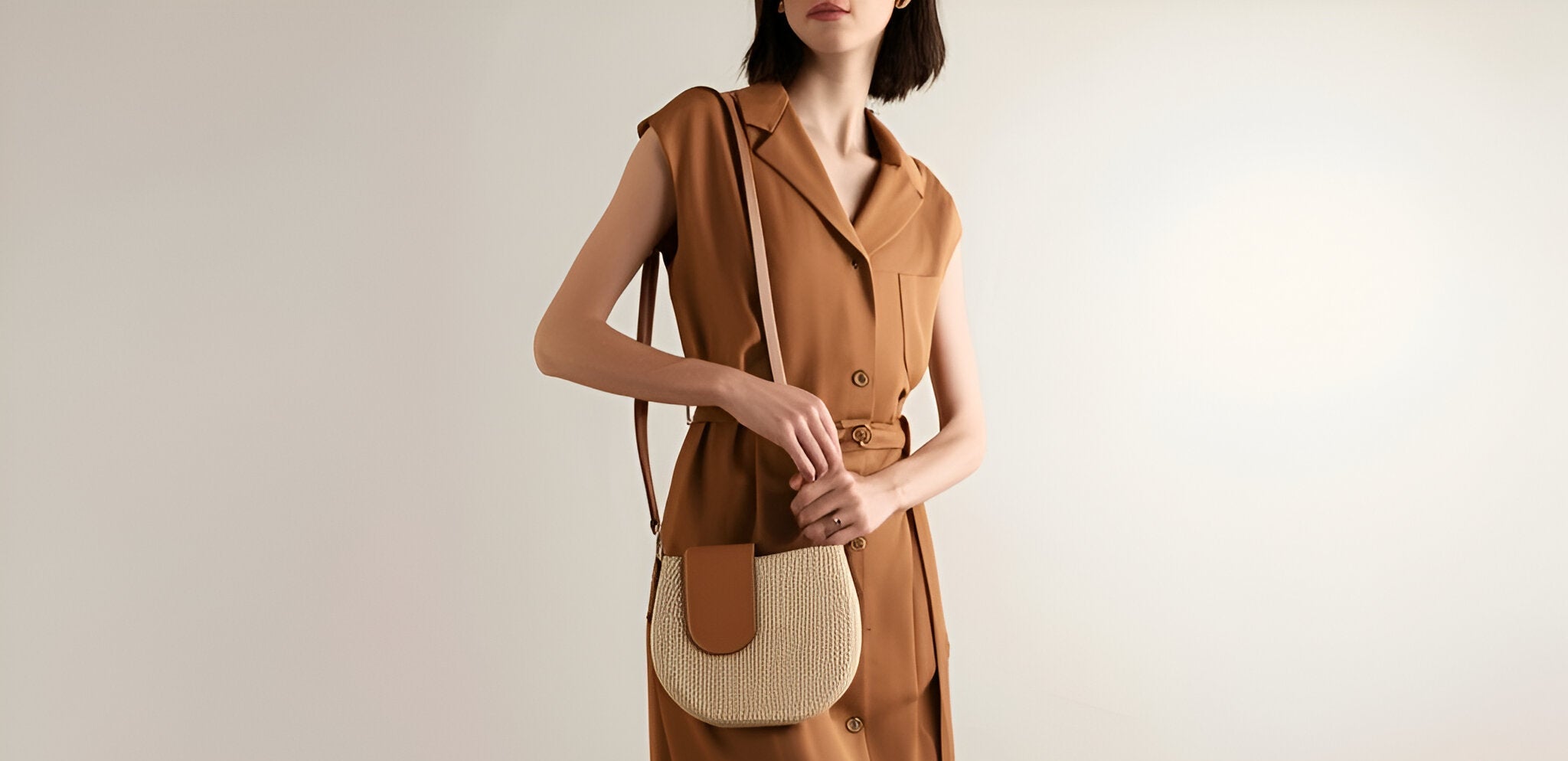 Stylish woman in sleeveless brown dress carrying a woven crossbody bag with tan leather flap against a neutral background.